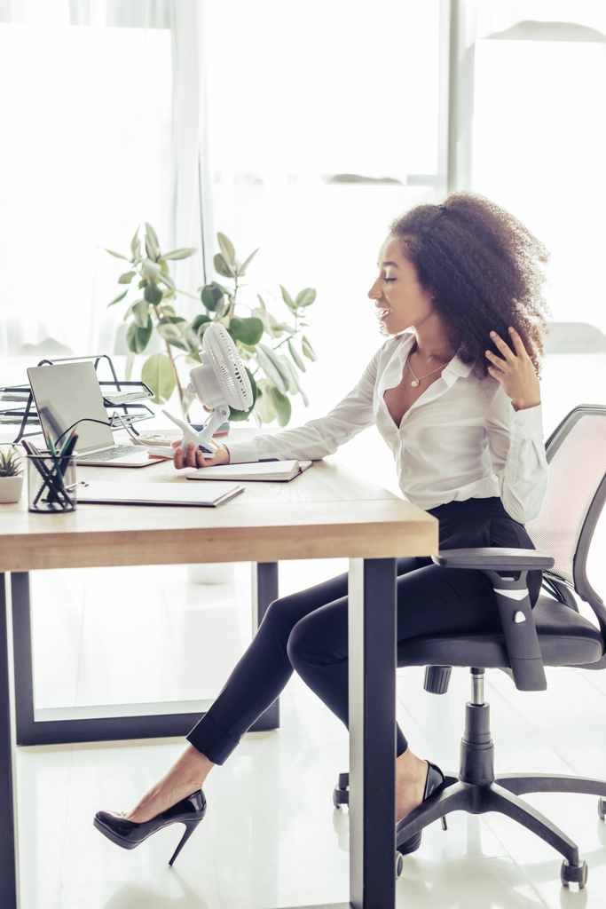 atraktivní africká americká žena, která sedí na pracovišti a drží elektrický ventilátor a současně trpí teplem v kanceláři - Fotografie, Obrázek
