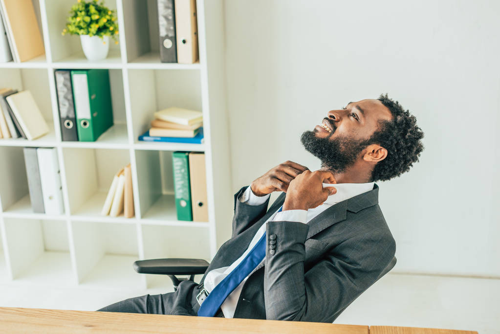 εξαντλημένος Αφρικανός Αμερικανός επιχειρηματίας κοιτάζοντας πάνω και αγγίζοντας γραβάτα, ενώ υποφέρουν από τη θερμότητα στο γραφείο - Φωτογραφία, εικόνα