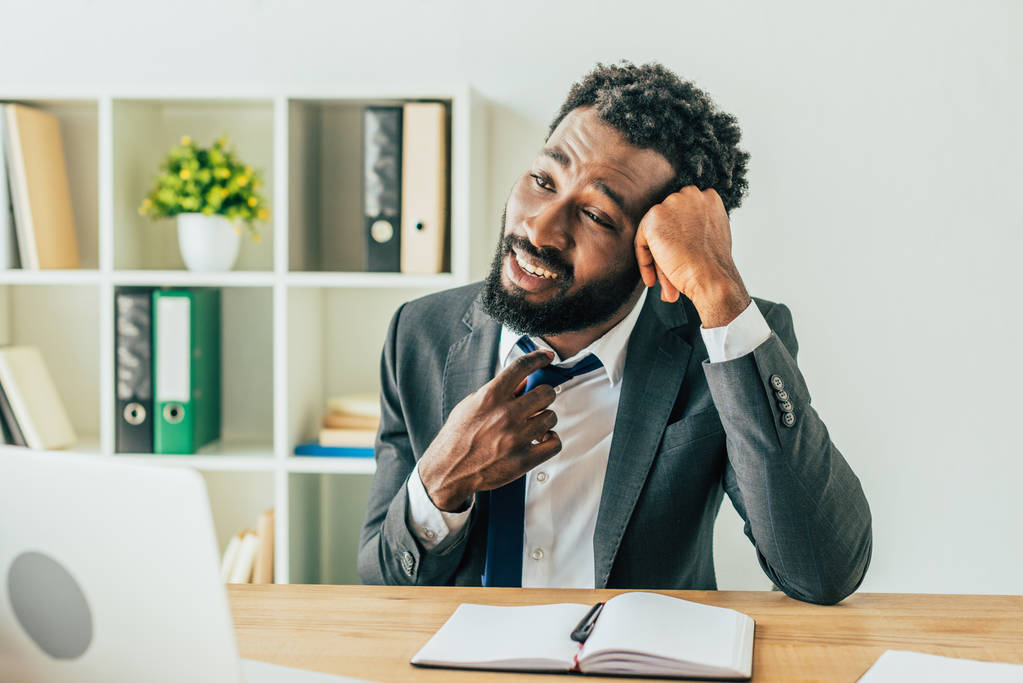 δυσαρεστημένος Αφρικανός Αμερικανός επιχειρηματίας αγγίζοντας γραβάτα, ενώ κάθονται στο χώρο εργασίας και υποφέρουν από τη θερμότητα στο γραφείο - Φωτογραφία, εικόνα