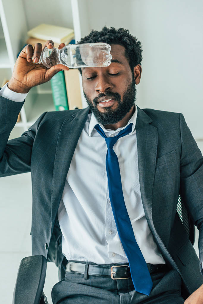 hombre de negocios afroamericano sosteniendo botella de plástico con agua cerca de la cabeza mientras sufre de calor en la oficina
 - Foto, imagen