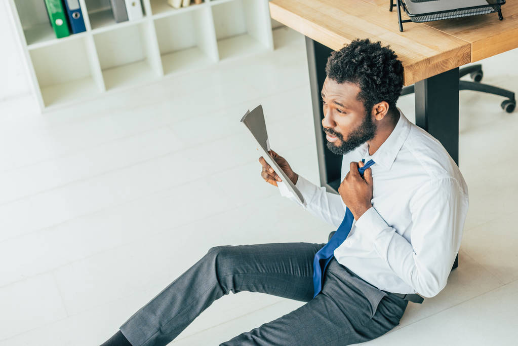 若いアフリカ系アメリカ人のビジネスマンが床に座り、オフィスの暑さに苦しみながらフォルダで手を振っている - 写真・画像