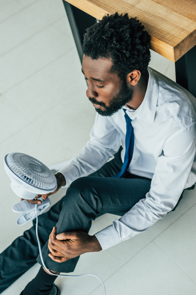 εναέρια θέα του αφρικανικού Αμερικανού επιχειρηματία που κάθεται στο πάτωμα με ηλεκτρικό ανεμιστήρα και υποφέρει από θερμότητα - Φωτογραφία, εικόνα