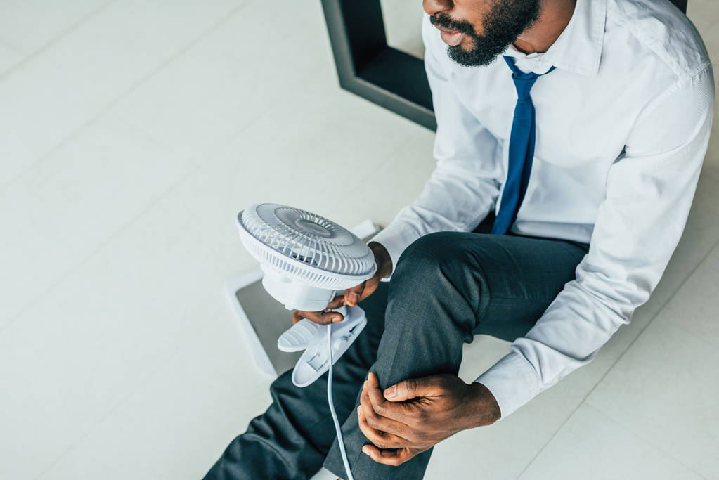 oříznutý pohled afroamerického podnikatele, který sedí na podlaze a drží v ruce foukající elektrický ventilátor  - Fotografie, Obrázek