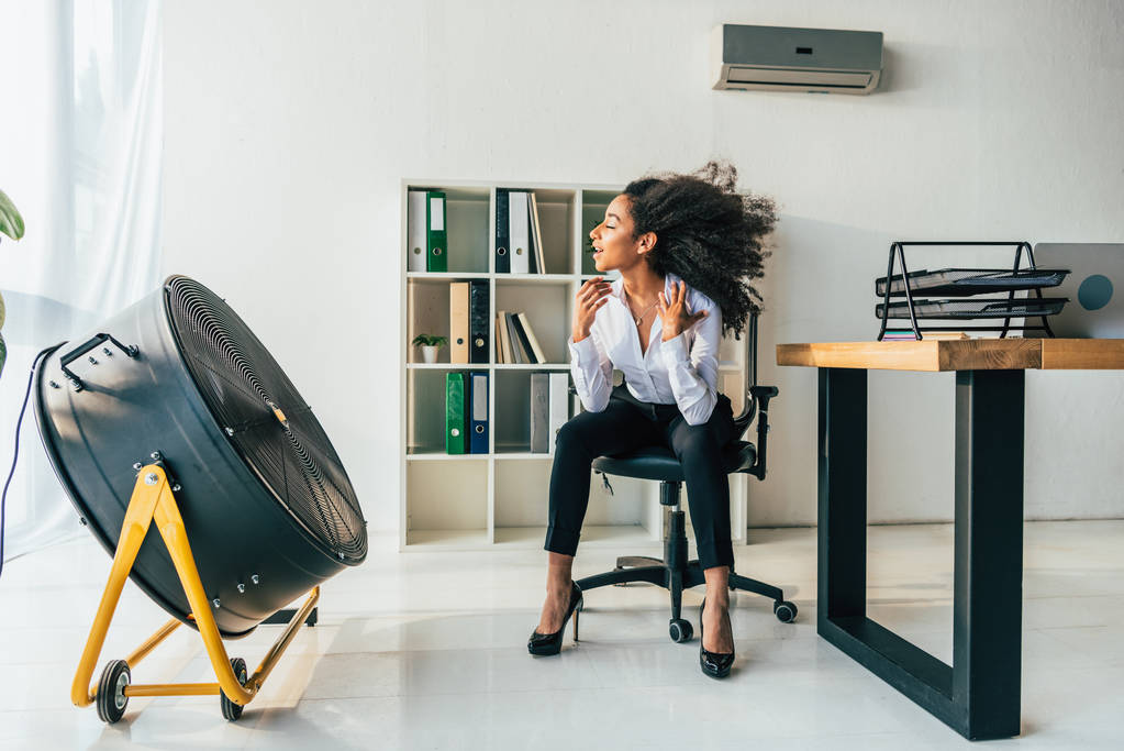 bonita mulher de negócios afro-americana sentada na cadeira de escritório na frente de soprar ventilador elétrico
 - Foto, Imagem