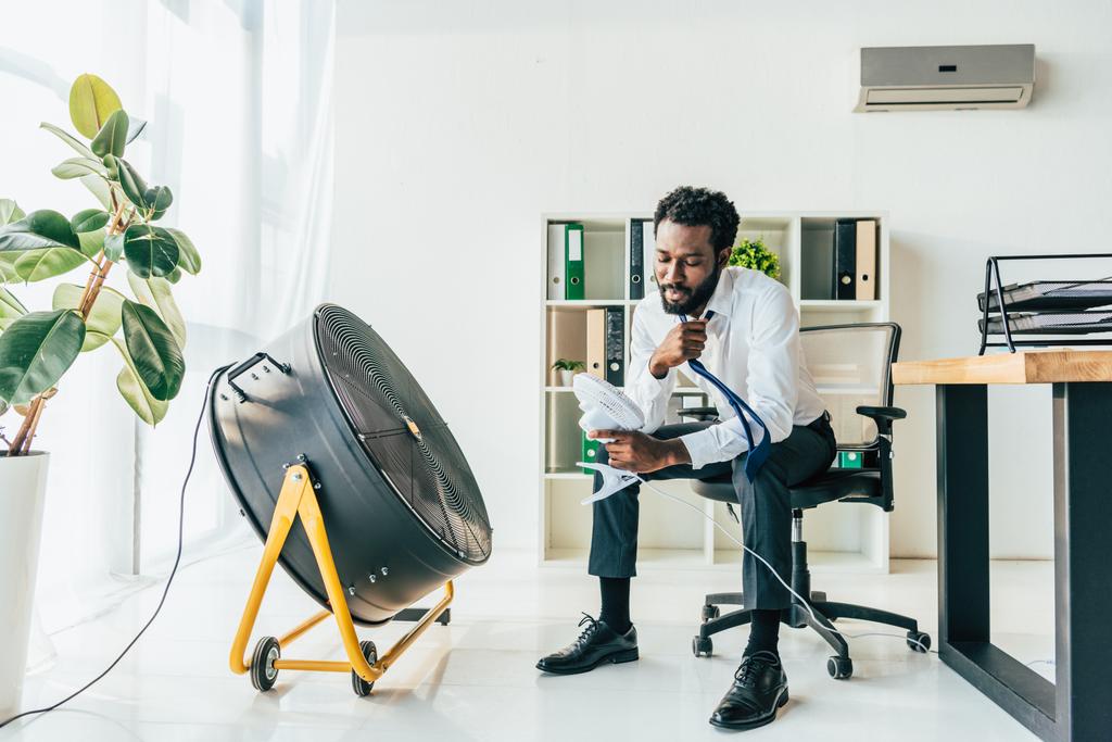 hombre de negocios afroamericano sosteniendo ventilador de escritorio mientras está sentado frente al ventilador eléctrico en la oficina
 - Foto, Imagen