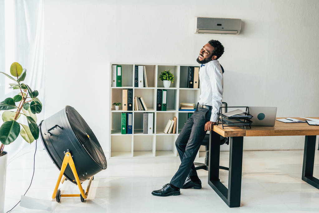 uitgeput Afro-Amerikaanse zakenman staande voor elektrische ventilator in kantoor - Foto, afbeelding