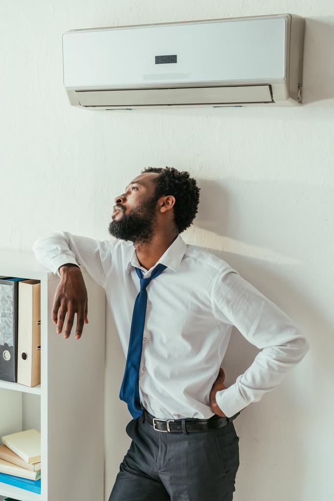思慮深いアフリカ系アメリカ人のビジネスマンは、エアコンの下に立って、オフィスで腰に手を握っている - 写真・画像