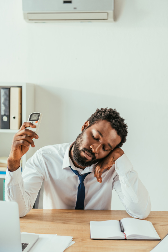 疲れ果てたアフリカ系アメリカ人のビジネスマンは、目を閉じて座って、エアコンのリモコンを保持しています - 写真・画像