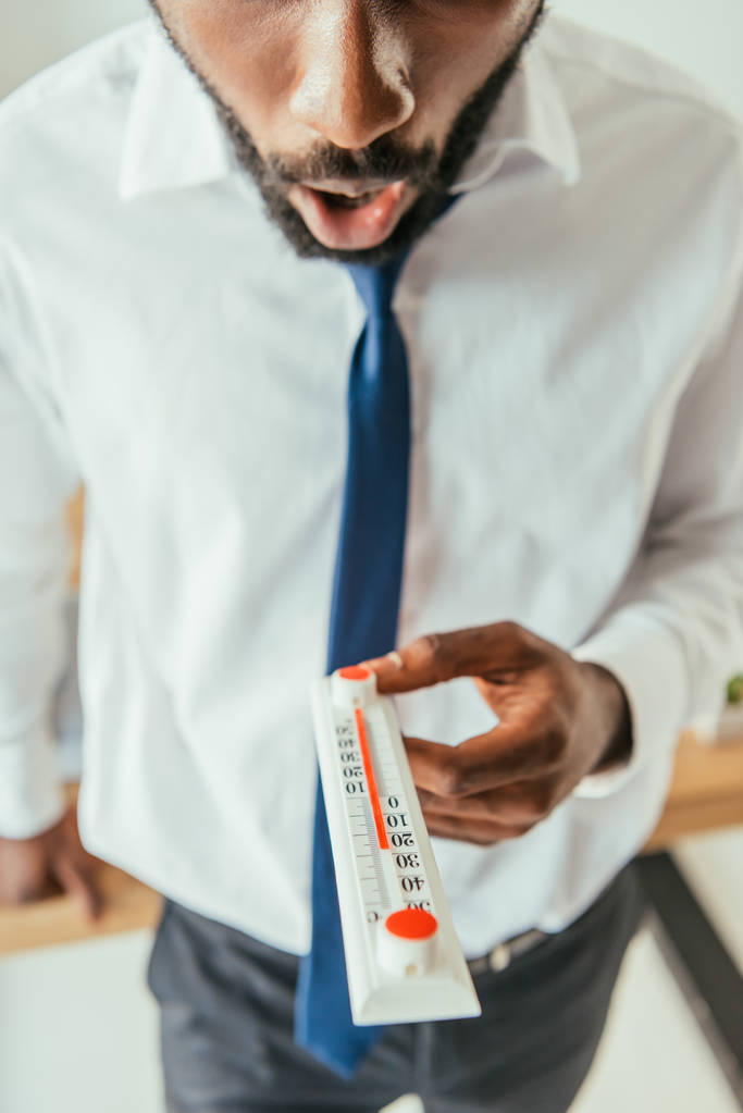 Ausgeschnittene Ansicht eines schockierten afrikanisch-amerikanischen Geschäftsmannes mit Thermometer, das hohe Temperaturen im Büro anzeigt - Foto, Bild