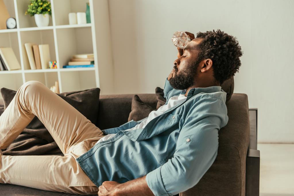 Erschöpfter afrikanisch-amerikanischer Mann liegt mit geschlossenen Augen auf Couch und hält Flasche mit Wasser in der Nähe des Kopfes, während er unter Hitze leidet - Foto, Bild
