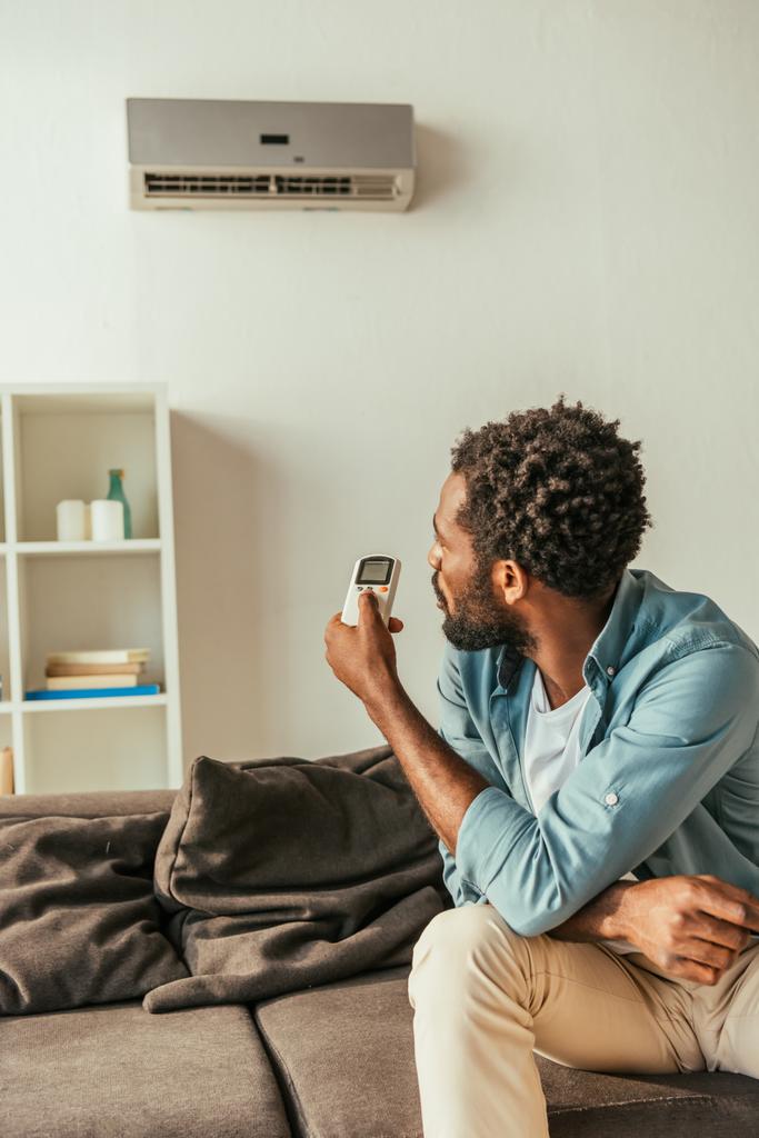 African American man met Air Conditioner afstandsbediening terwijl zittend op de Bank thuis - Foto, afbeelding