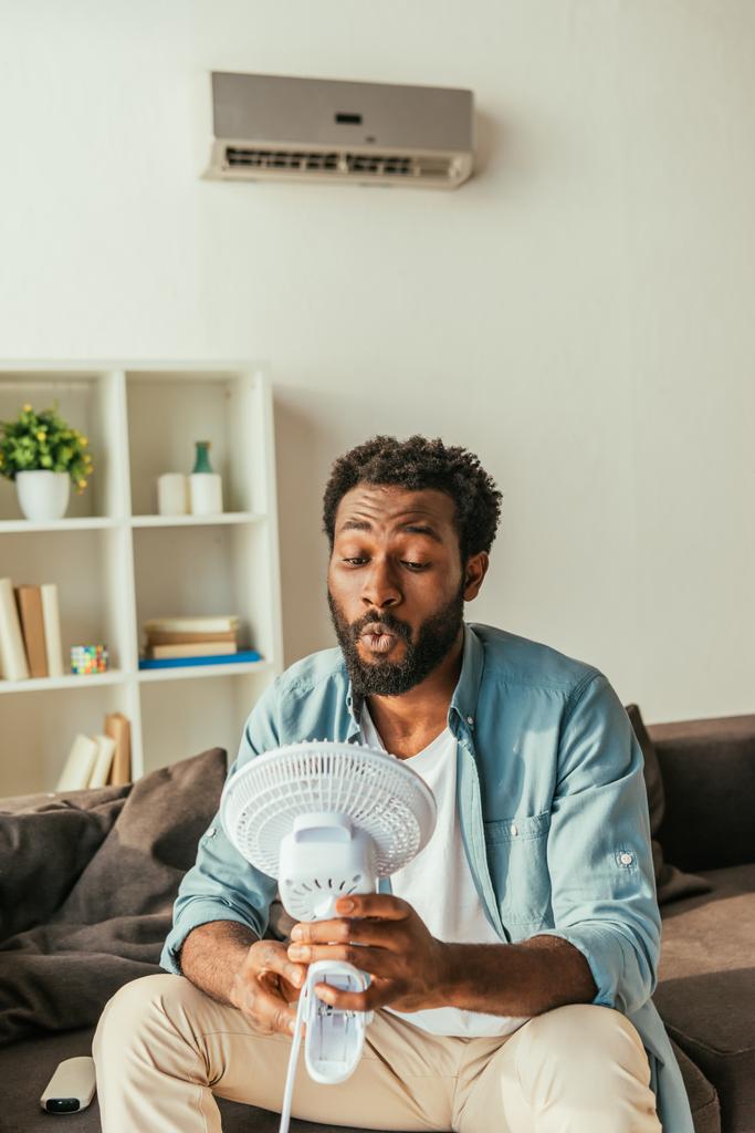 красивий афроамериканський чоловік тримає дме електричний вентилятор, сидячи на дивані і страждаючи від спеки вдома
 - Фото, зображення