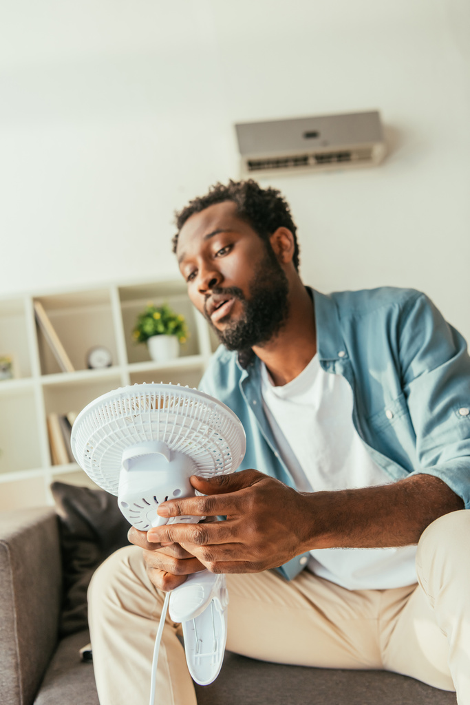 ソファに座り、自宅で暑さに苦しんでいる間、扇風機を吹いている疲れ果てたアフリカ系アメリカ人の男性 - 写真・画像