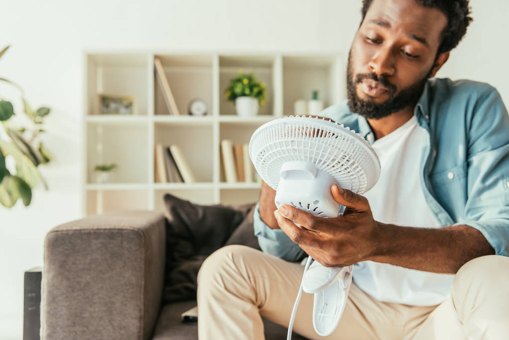измученный африканский американец держит дующий электрический вентилятор в то время как страдает от жары дома
 - Фото, изображение