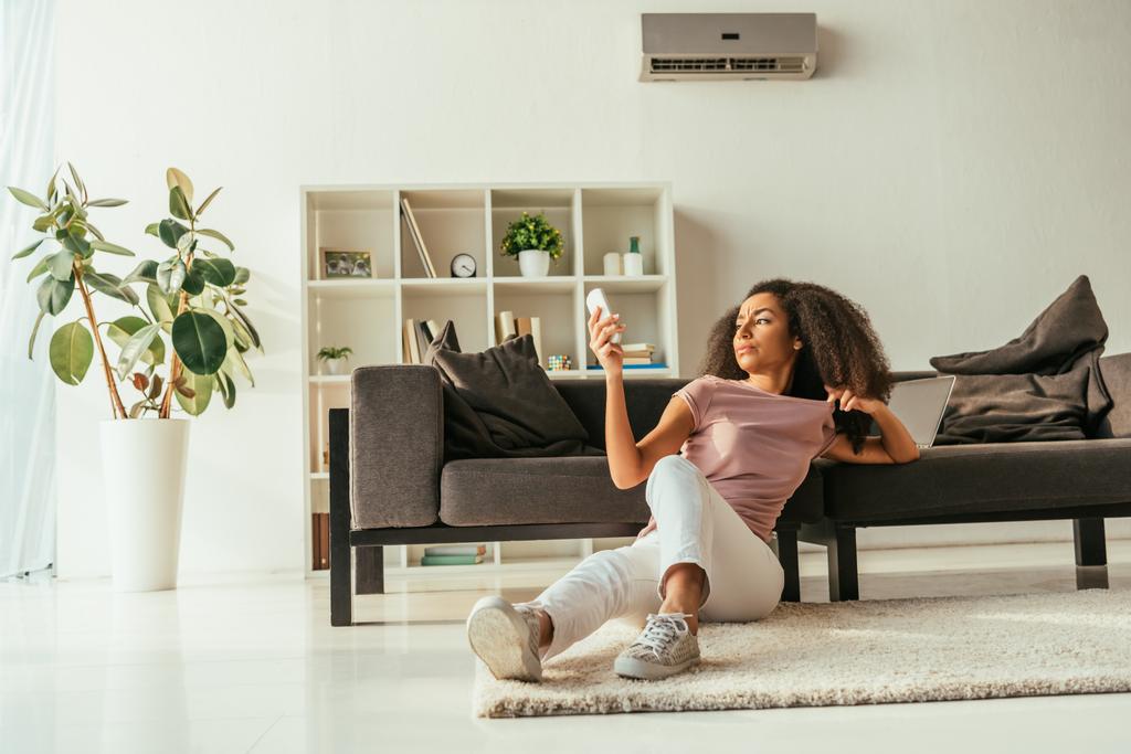 attraktive afrikanisch-amerikanische Frau sitzt auf dem Boden und benutzt eine Fernbedienung der Klimaanlage, während sie zu Hause unter der Sommerhitze leidet - Foto, Bild