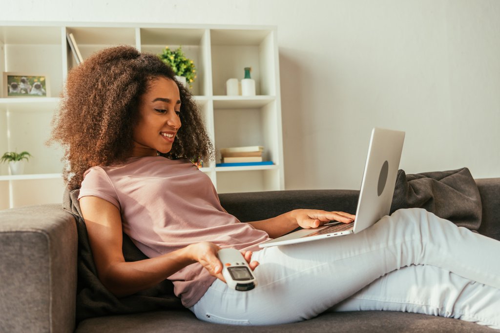 usmívající se africká americká žena pomocí laptopu a přidržování dálkového ovladače klimatizace při lhaní na pohovce a používání přenosného počítače - Fotografie, Obrázek