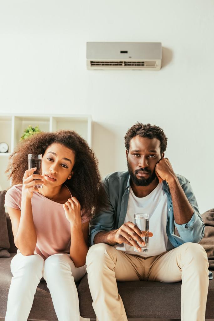unhappy african american man and woman sitting on sofa and drinking water while suffering from summer heat at home - Photo, Image