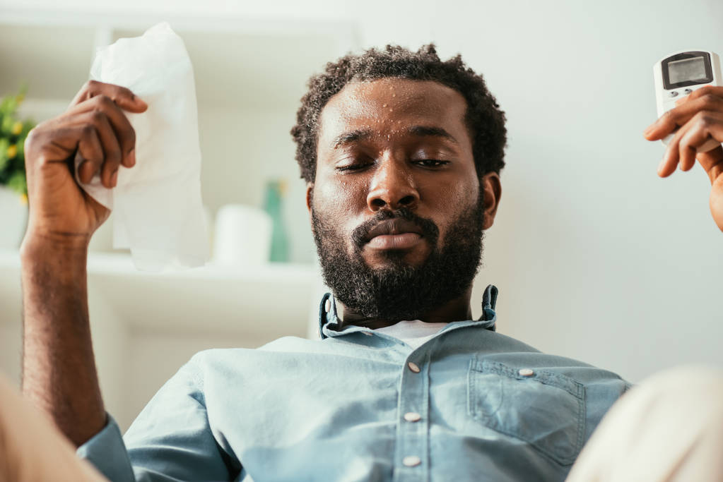African American man met zweterige gezicht Holding servet en afstandsbediening terwijl het lijden van warmte thuis - Foto, afbeelding