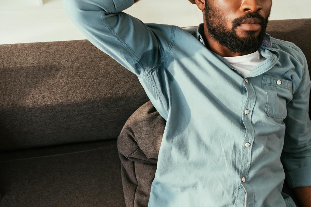 vista parziale dell'uomo afroamericano in camicia sudata seduto sul divano e affetto da calore
 - Foto, immagini