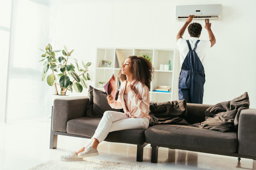 mujer afroamericana bonita sentada en el sofá y saludando con la revista, mientras que el reparador afroamericano fija el aire acondicionado
 - Foto, imagen