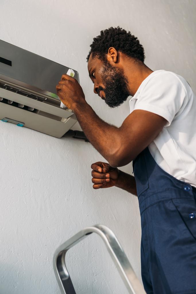 pozorný africký americký opravák stojící na žebříku a upevňukní klimatizaci - Fotografie, Obrázek