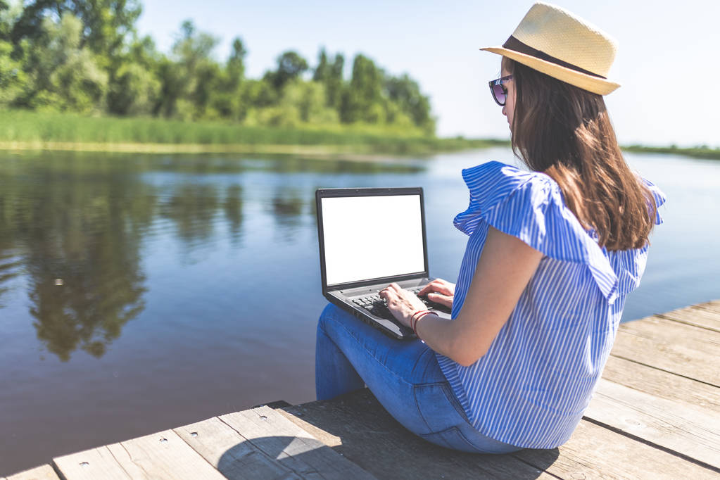 Side view of young woman sitting on pier and using blank screen laptop. Nature and technology concepts. - Photo, Image