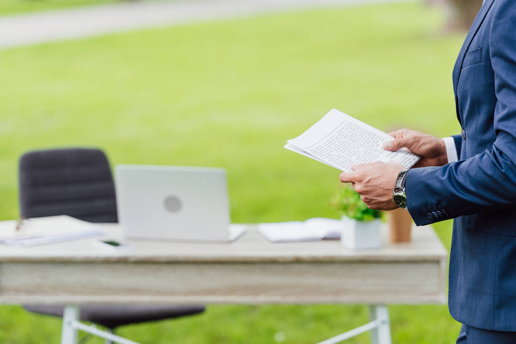 cropped view of young businessman holding newspaper and standing in park near table - Photo, Image