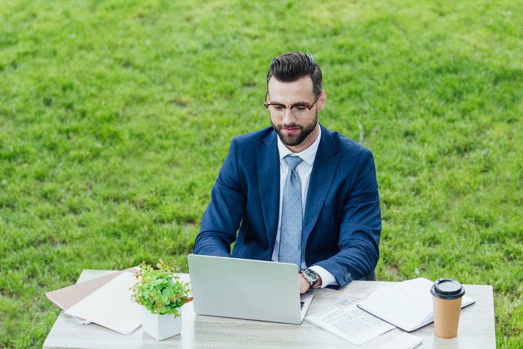 νέος επιχειρηματίας σε τυπική φθορά χρησιμοποιώντας φορητό υπολογιστή, ενώ κάθονται στο πάρκο πίσω από το λευκό γραφείο τραπέζι  - Φωτογραφία, εικόνα