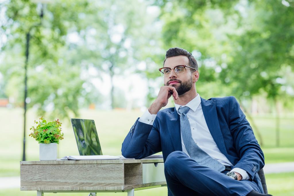 молодий бізнесмен сидить за столом з ноутбуком і рослиною в парку, дивлячись далеко
 - Фото, зображення