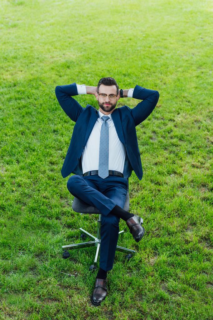 высокий угол зрения бизнесмена, сидящего в офисном кресле в парке со скрещенными лагами и руками за головой
 - Фото, изображение