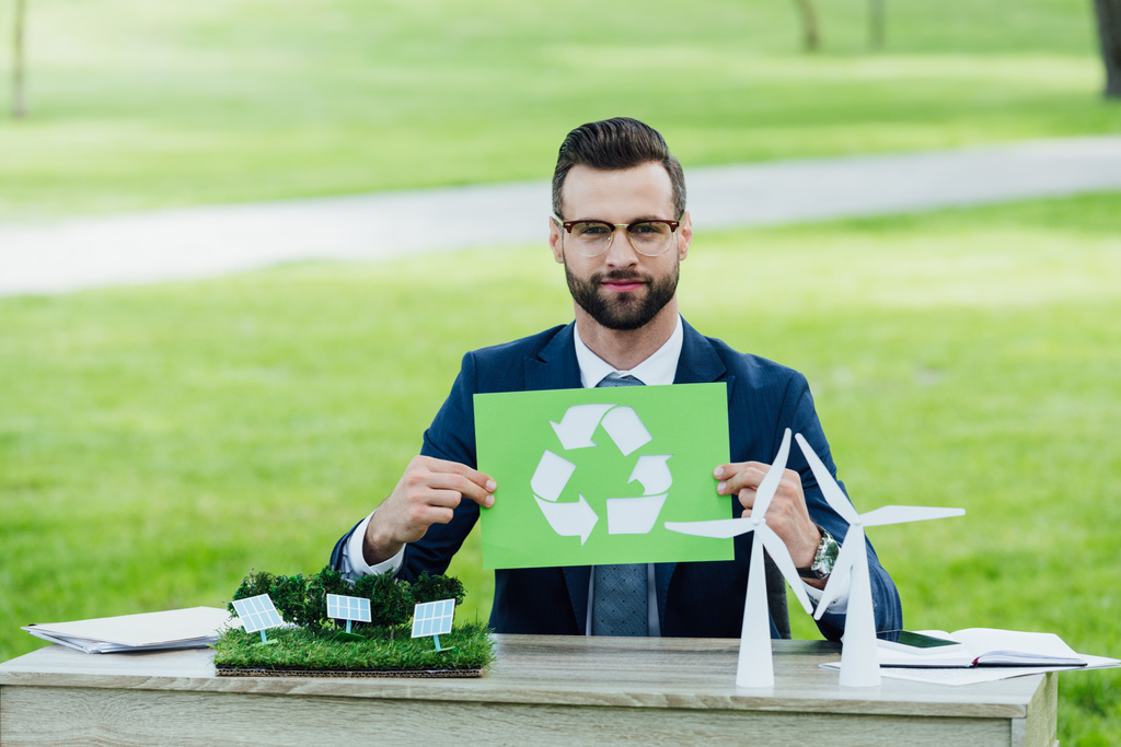 joven hombre de negocios en la mesa con molinos de viento y diseños de baterías solares con tarjeta de retención con inscripción de reciclaje
 - Foto, Imagen