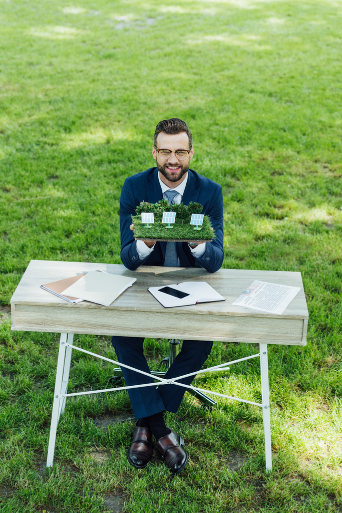 公園のオフィスチェアに座りながら、ノートパソコンやスマートフォンでテーブルの上に太陽電池でレイアウトを保持するビジネスマンの完全な長さのビュー  - 写真・画像