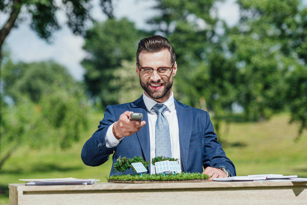 young businessman with glasses sitting at table with sun batteries layout, looking at camera, smiling and presenting control panel  - Photo, Image