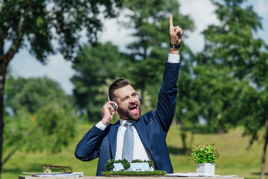 excited young businessman sitting at table with sun batteries layout and flowerpot, listening to music with headphones and putting hand in air - Photo, Image