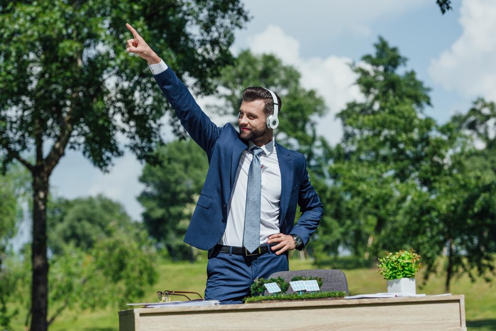 mladý podnikatel poslouchá hudbu ve sluchátkách a tančí v parku u stolu s rozložením slunečních baterií a květináčem - Fotografie, Obrázek