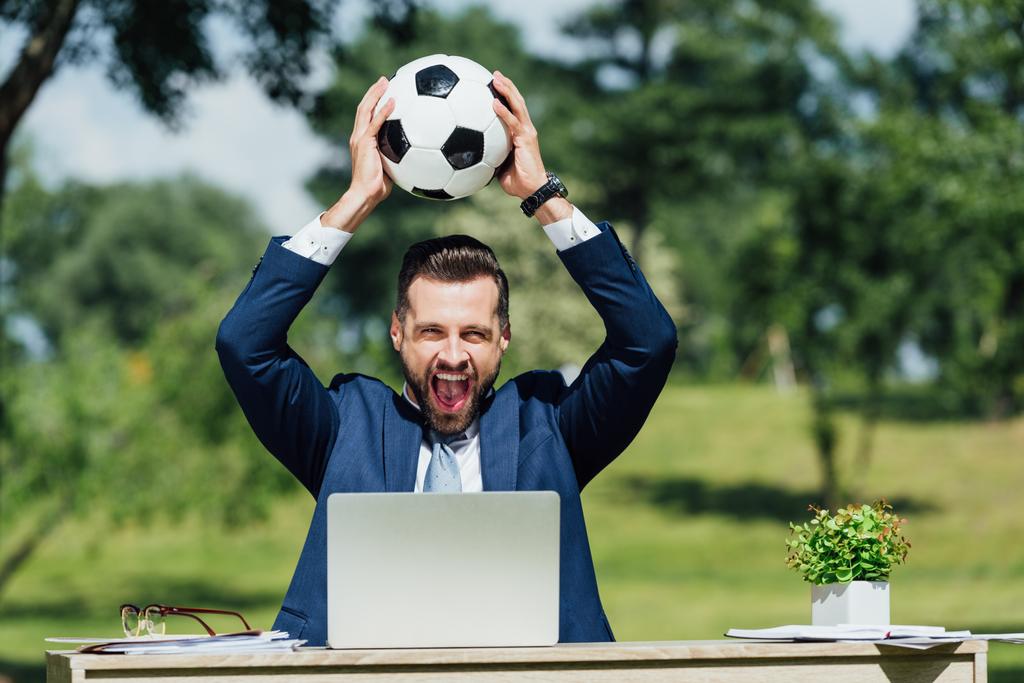jeune homme d'affaires assis à table avec ordinateur portable, pot de fleurs et lunettes, souriant et tenant le football au-dessus de la tête
 - Photo, image