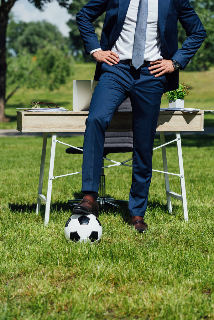 Ausgeschnittene Ansicht eines Geschäftsmannes, der mit einem Bein auf einem Fußballball neben einem Tisch im Park steht und die Hände auf die Hüften legt - Foto, Bild