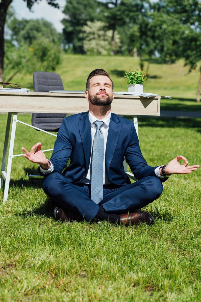 公園のテーブルの近くの草の上に座って瞑想するビジネスマン  - 写真・画像