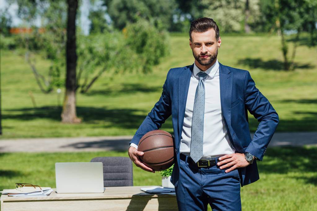 homem de negócios em pé mesa limpa no parque, segurando basquete e olhando para a câmera
 - Foto, Imagem