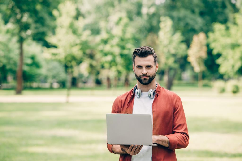 молодой человек, стоящий в парке с ноутбуком в руках и смотрящий в камеру
 - Фото, изображение