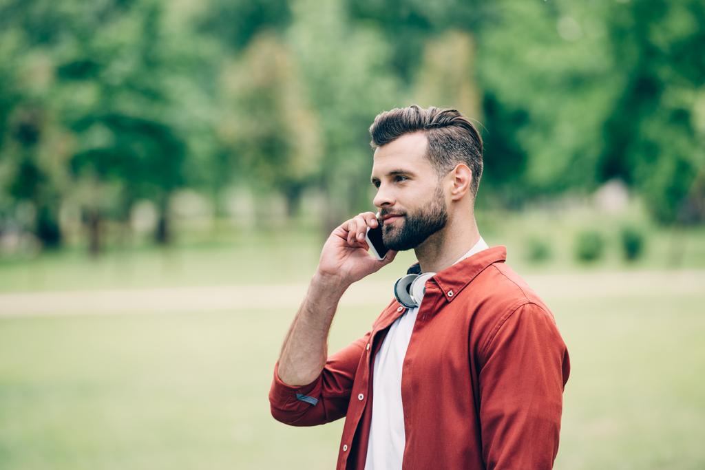 νεαρός άντρας στέκεται στο πάρκο και μιλάει στο smartphone  - Φωτογραφία, εικόνα