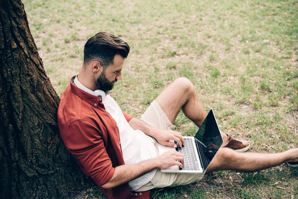 Seitenansicht eines jungen Mannes, der auf Gras neben Baum sitzt und Laptop benutzt - Foto, Bild