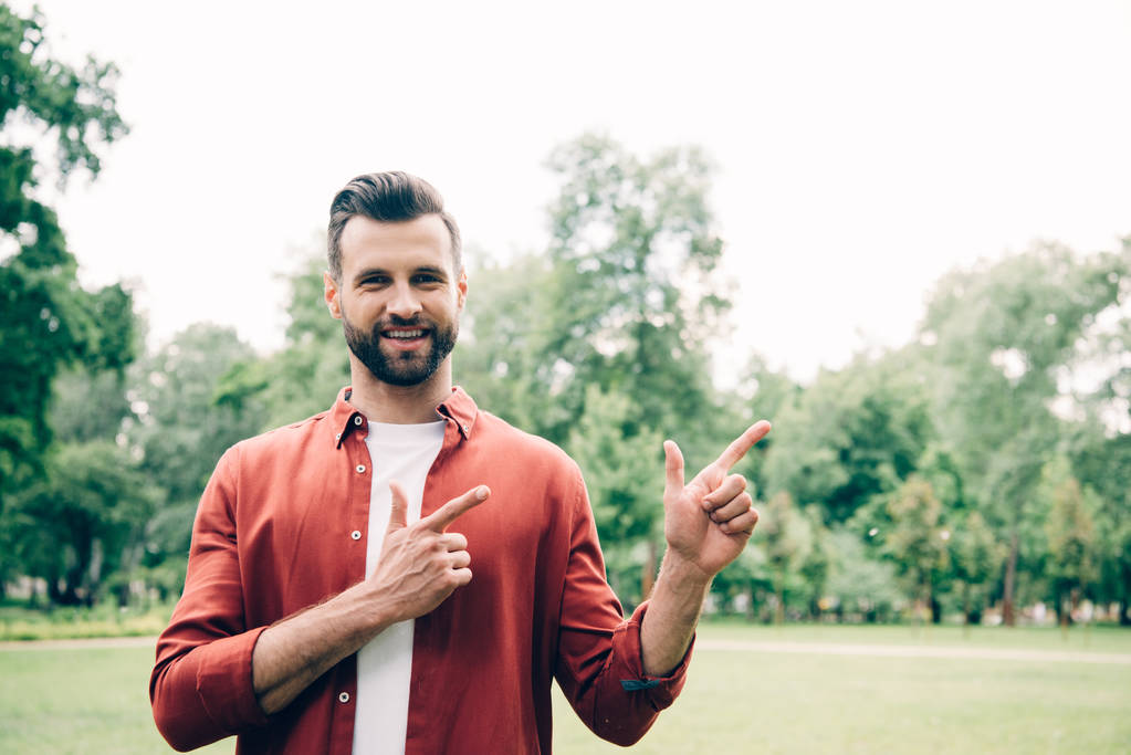 człowiek w czerwonej koszulce stojącej w parku, patrząc na kamerę i wskazując palcami  - Zdjęcie, obraz