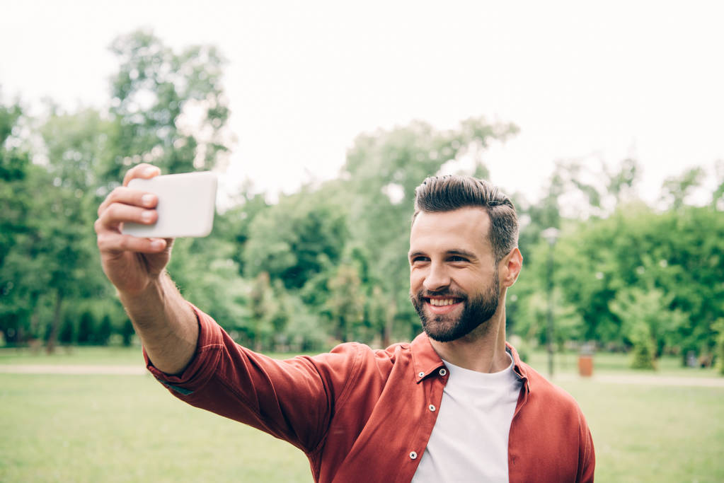 przystojny człowiek stoi w parku, uśmiechając się i biorąc selfie  - Zdjęcie, obraz