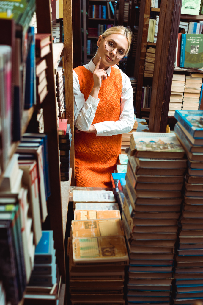 mooie en blonde vrouw in glazen en oranje jurk op zoek weg in de bibliotheek   - Foto, afbeelding