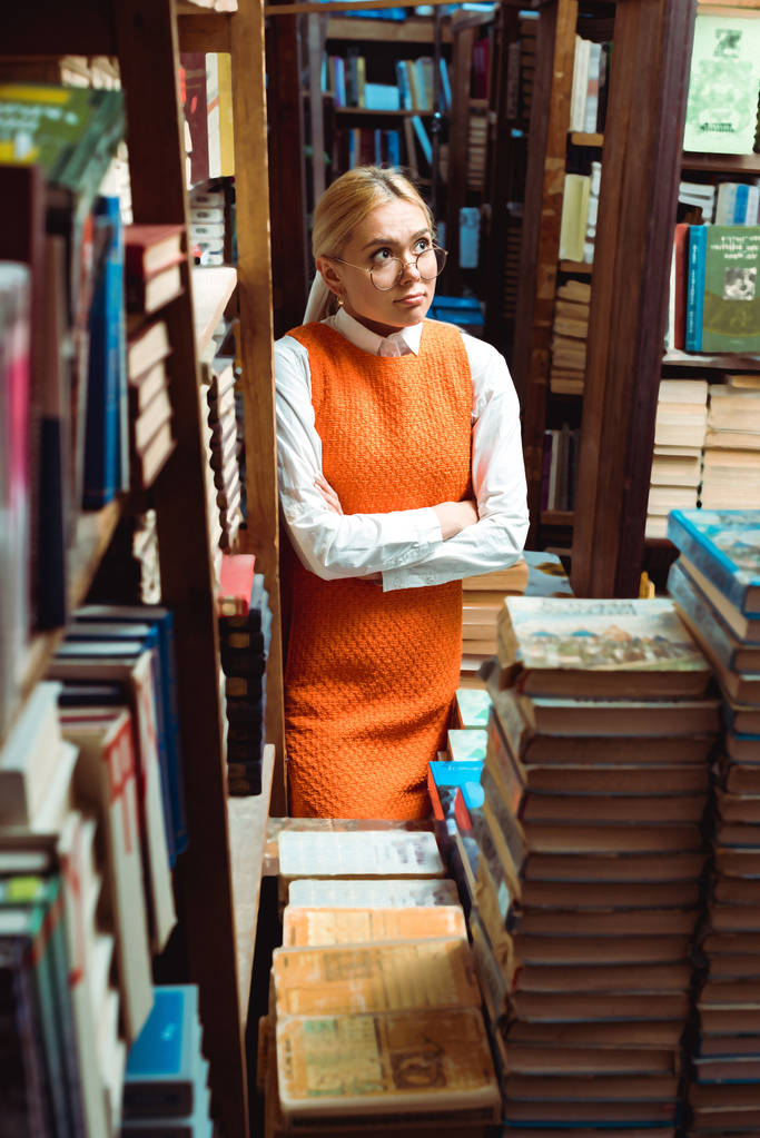 szép és szomorú nő a szemüveg és narancssárga ruhában a keresztkaron elnézett a könyvtárban   - Fotó, kép