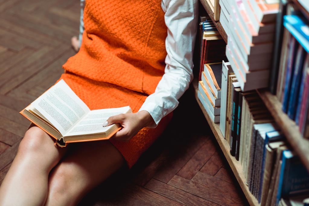 木製の床に座って本を保持しているオレンジ色のドレスを着た女性のトリミングされたビュー  - 写真・画像