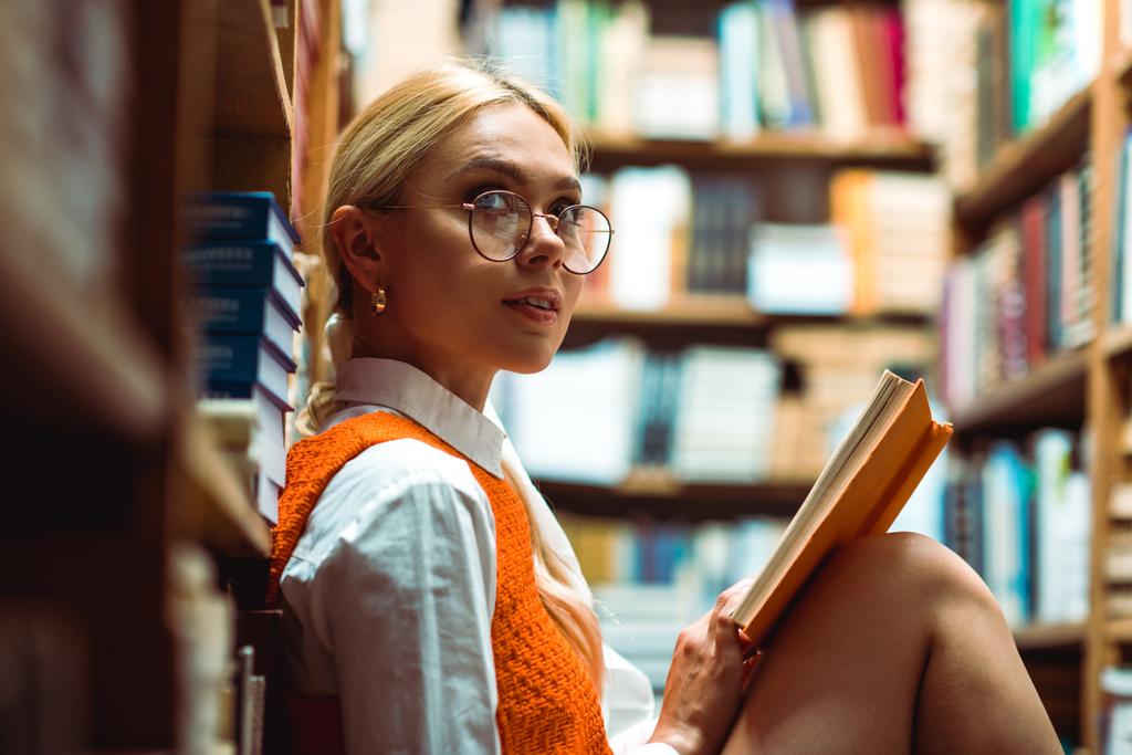 schöne und blonde Frau mit Brille, Buch in der Hand und wegschauend in der Bibliothek  - Foto, Bild