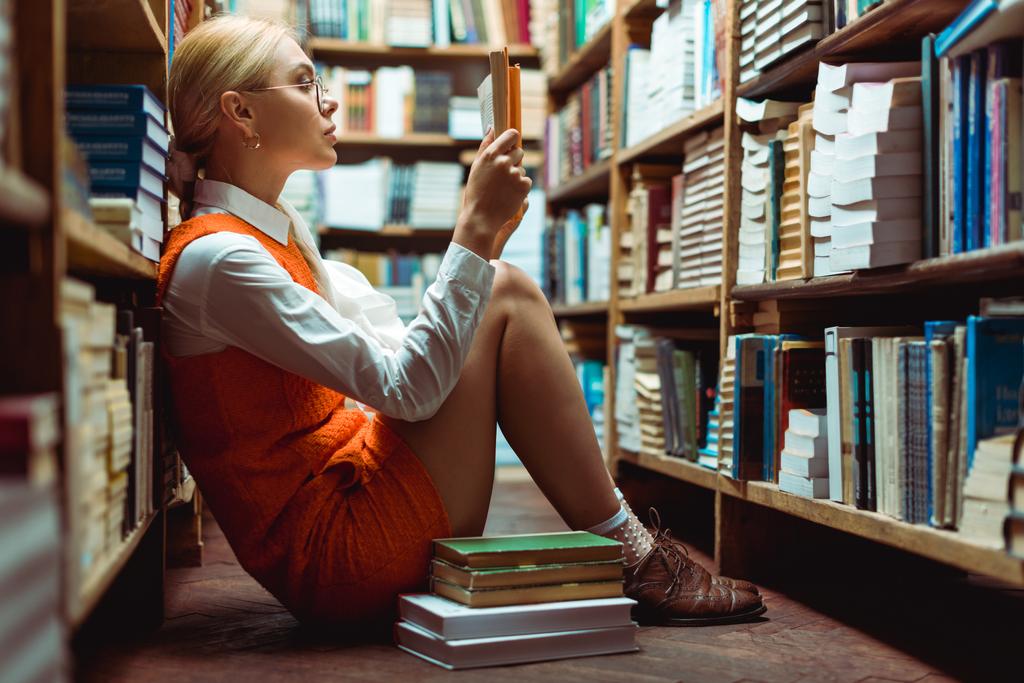 όμορφη και ξανθιά γυναίκα σε γυαλιά διαβάζοντας βιβλίο και καθισμένος στο πάτωμα στη βιβλιοθήκη  - Φωτογραφία, εικόνα