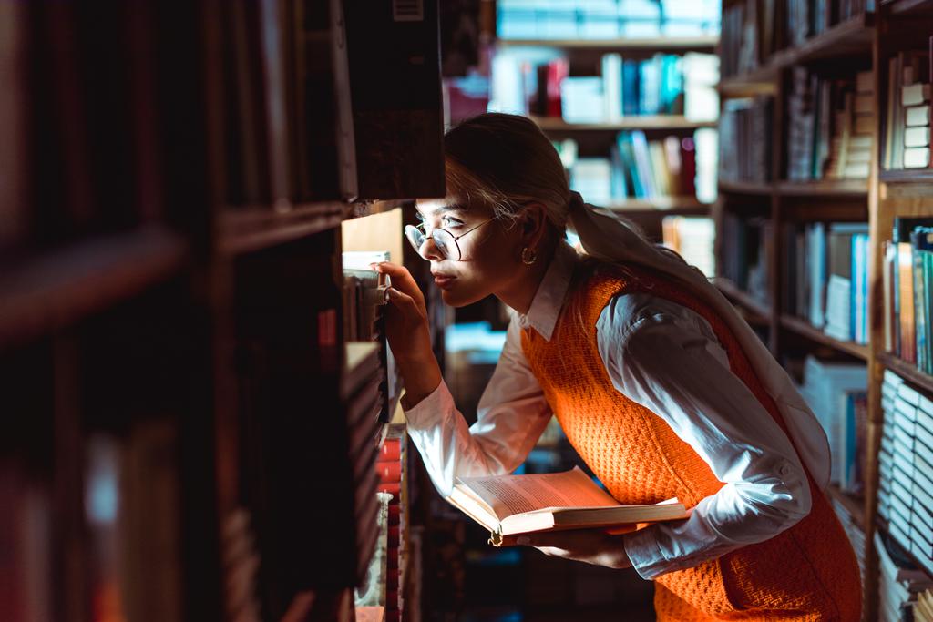 όμορφη γυναίκα με γυαλιά κοιτάζοντας μακριά και κρατώντας το βιβλίο στη βιβλιοθήκη  - Φωτογραφία, εικόνα